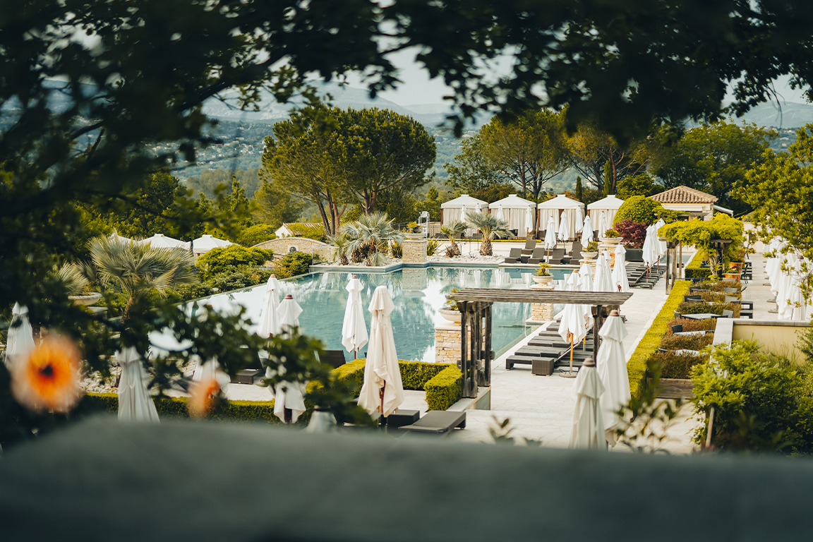 A large European swimming pool surrounded by sun loungers, umbrellas and manicured gardens