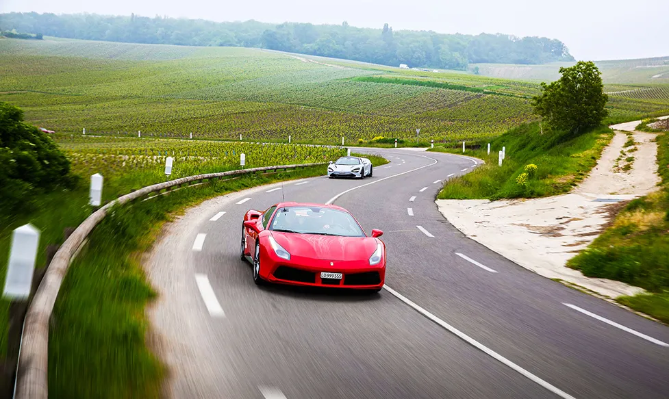 A red Ferrari 488 leads a white McLaren 720S through corners roads next to vineyards in Champagne, France