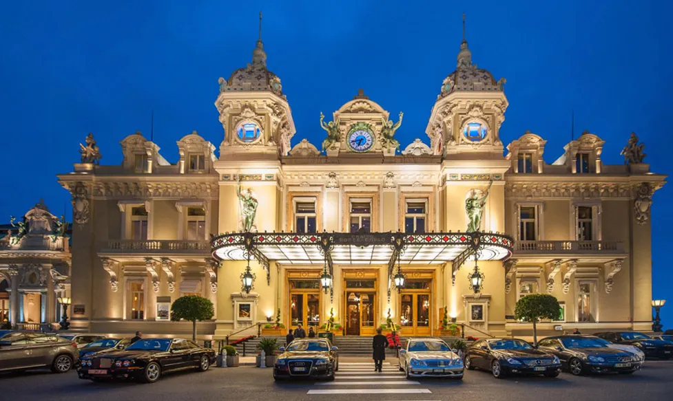 The iconic Casino du Monte Carlo is all lit up in front of a dark blue evening sky