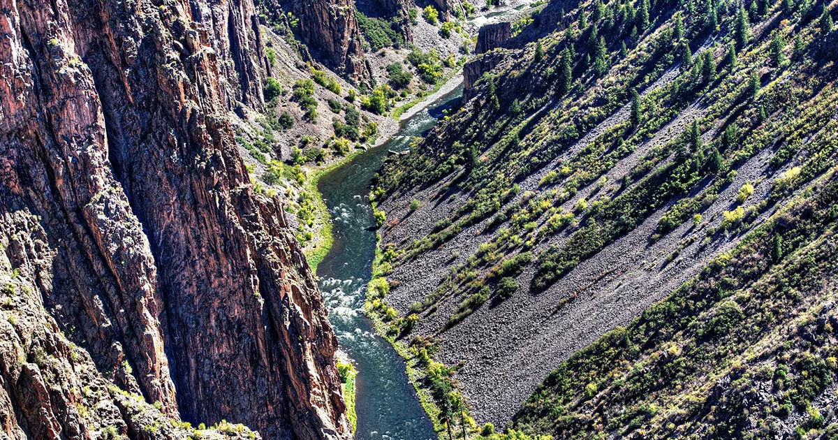 Steep banks of a canyon with bushes and trees descending to a fast flowing river.
