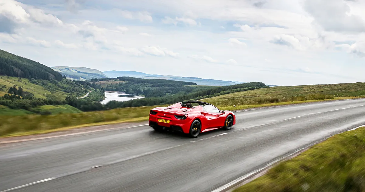 A red Ferrari cruises through rolling green hills in the English countryside.
