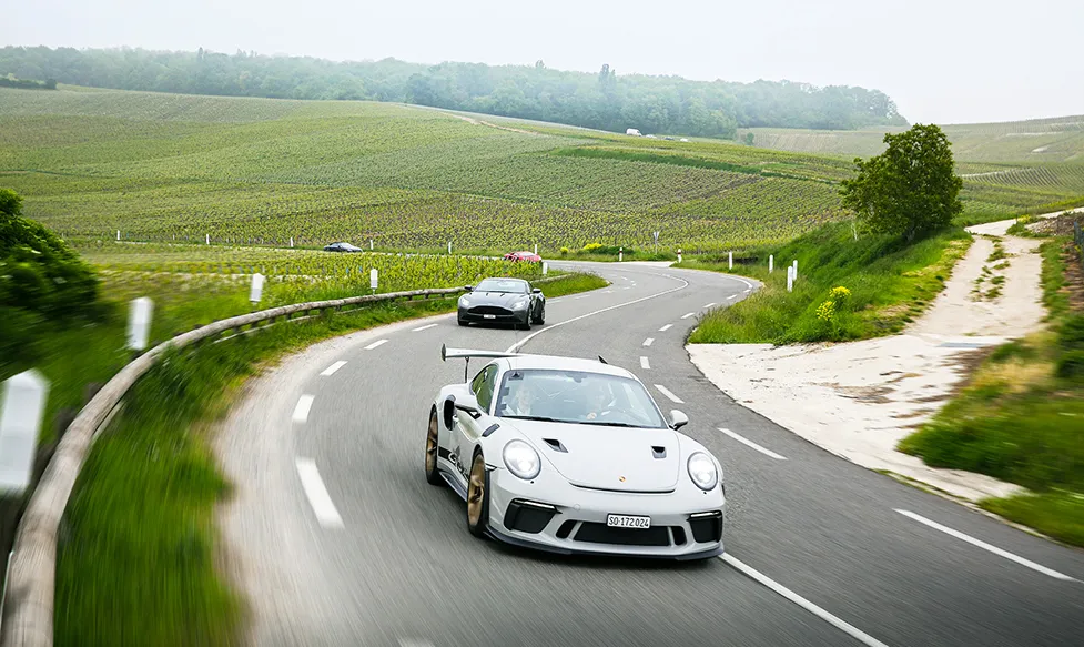 A grey Porsche 911 GT3 RS leads an Aston Martin through a sweeping section of corners flanked by vineyards in France