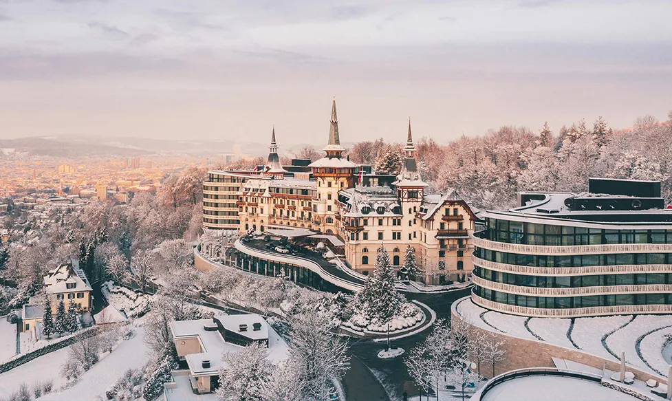 An exterior view of the Colder Grand Zurich hotel covered in snow as the sun rises
