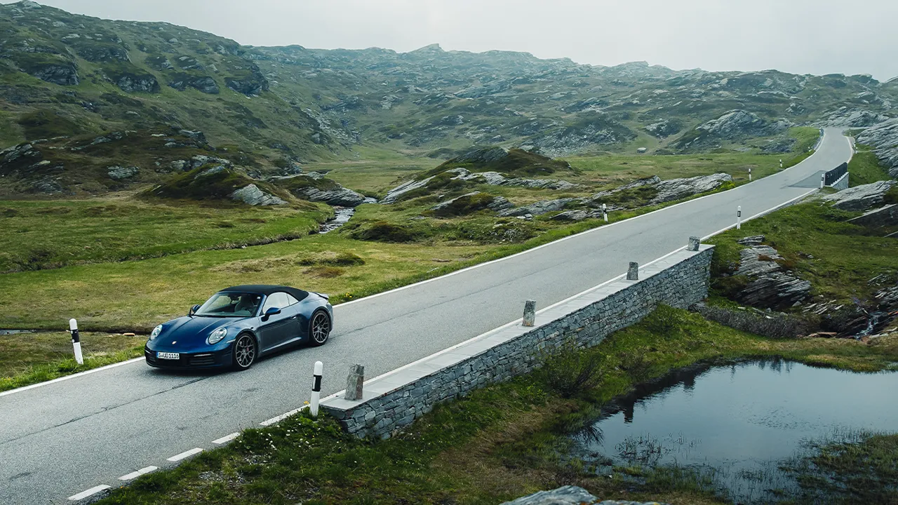 A blue Porsche convertible sportscar with a black hood cruises through a rugged and green country road on a misty morning