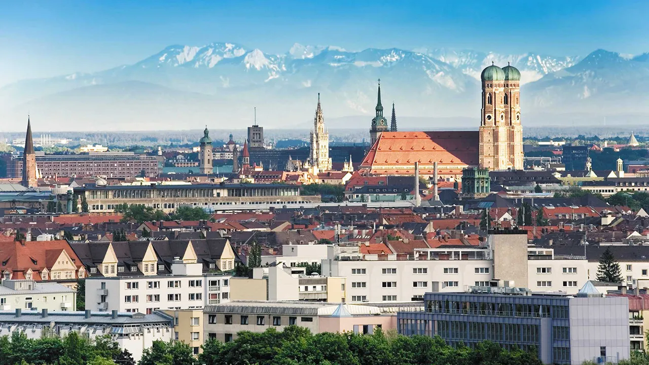 A backdrop of snowcapped mountains in Bavaria rise above low mist behind Munich's historic architecture